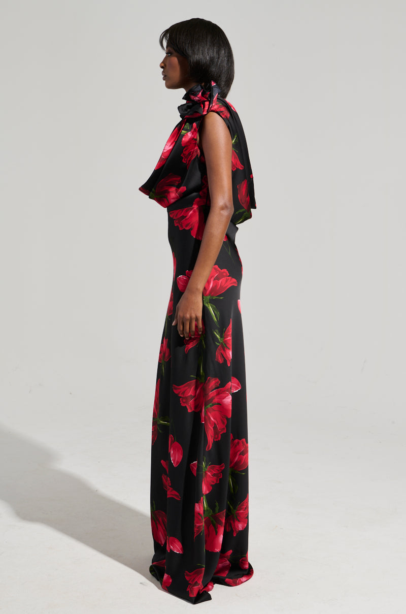 A person stands sideways, wearing the ROSE MAXI DRESS, which features a long, sleeveless silhouette adorned with vibrant red floral patterns. The satin fabric lends an elegant touch to the high-collared design, accentuated by a draped detail, all set against a plain white background.