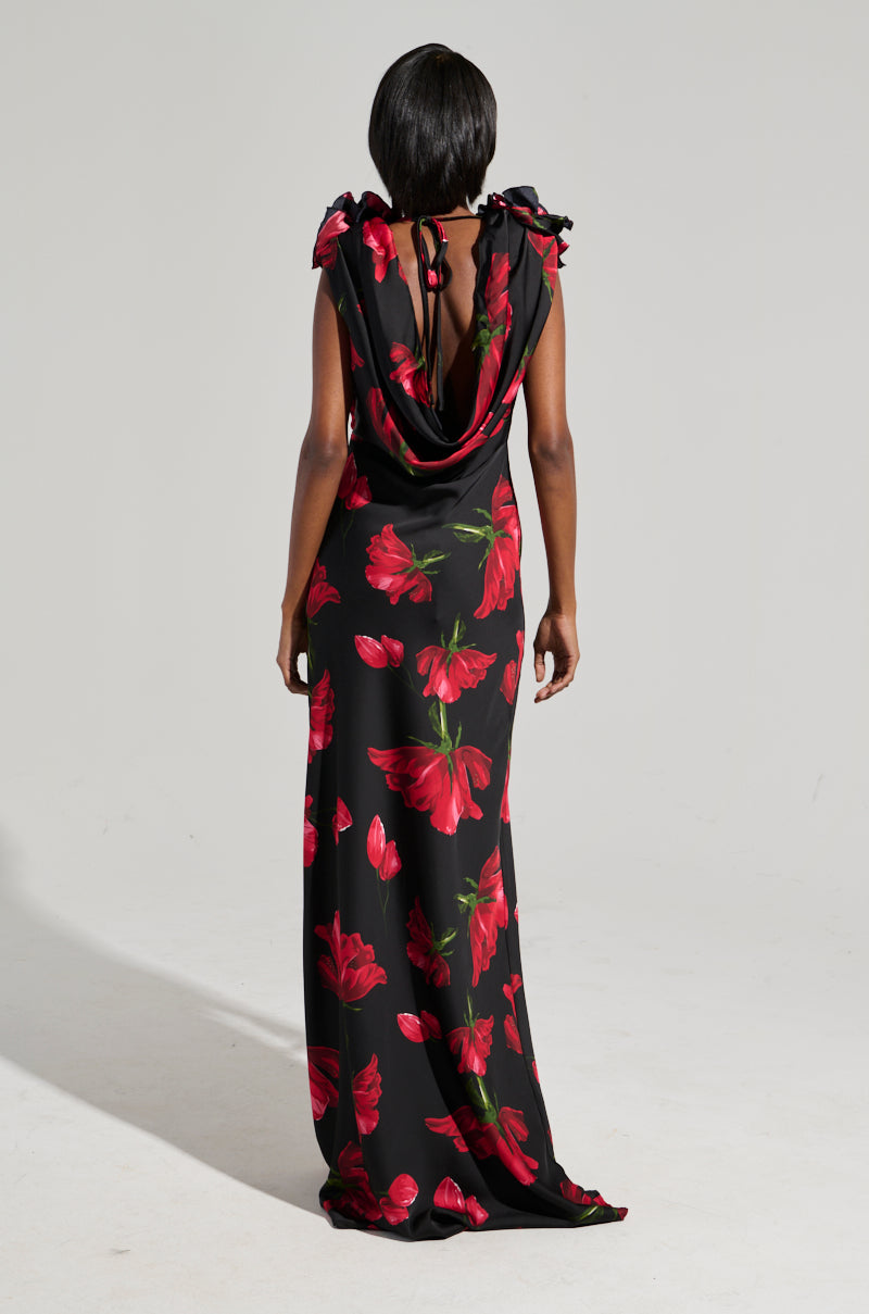 The image showcases the rear view of an individual in a ROSE MAXI DRESS, designed with luxurious satin fabric adorned with red floral patterns. This elegant dress is distinguished by its draped open back and ruffled shoulders, as the person stands with their hands resting by their sides against a plain backdrop.