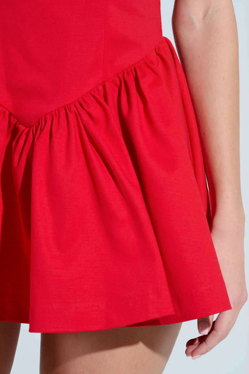 A close-up of a person in a vibrant Rosalia Stretch Linen Mini Dress in Red, featuring a ruffled, gathered waist and flared skirt. They stand against a plain light background, highlighting the dress's elegance.