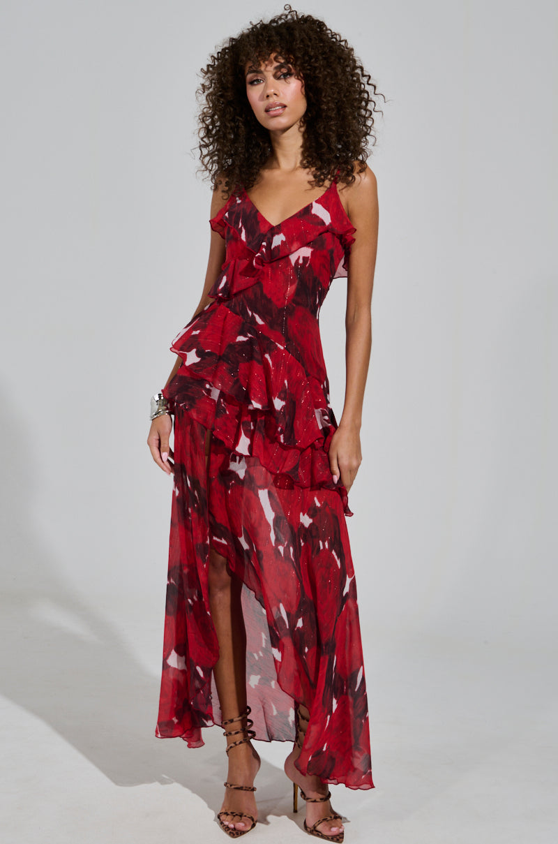 A woman with curly hair models the MAEVE PRINTED RUFFLE MAXI DRESS, highlighting its vibrant floral pattern and tiered ruffles. She effortlessly poses against a plain background in strappy heels, accessorized with a chic bracelet, showcasing the dress’s adjustable tank straps.