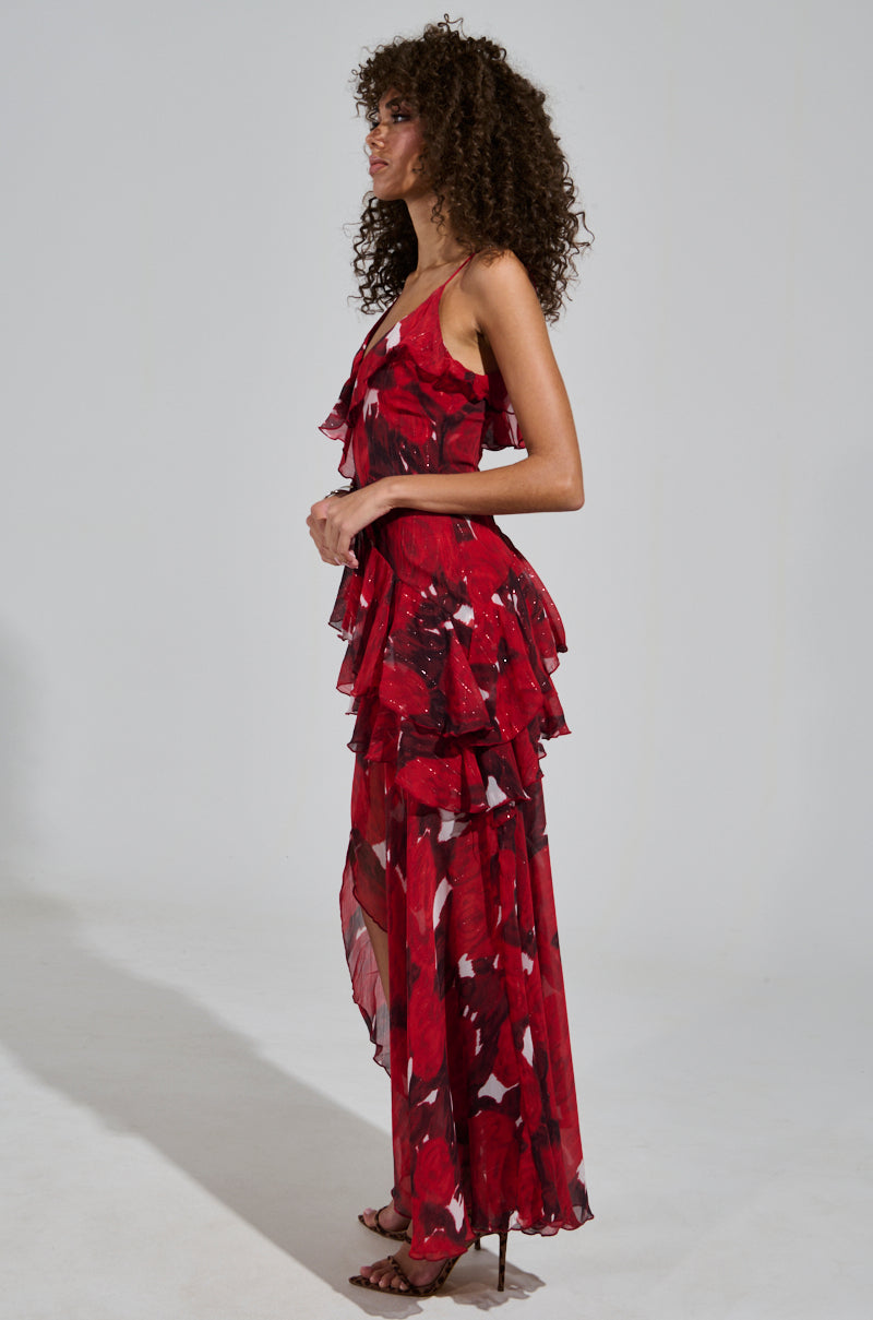 Wearing the MAEVE PRINTED RUFFLE MAXI DRESS, a person poses sideways in a light-colored, plain backdrop. The dress features a flowing red floral pattern with layered ruffles that beautifully accentuate their ensemble. Their curly hair adds to the elegance of their look, which is completed with high-heeled shoes and adjustable tank straps.