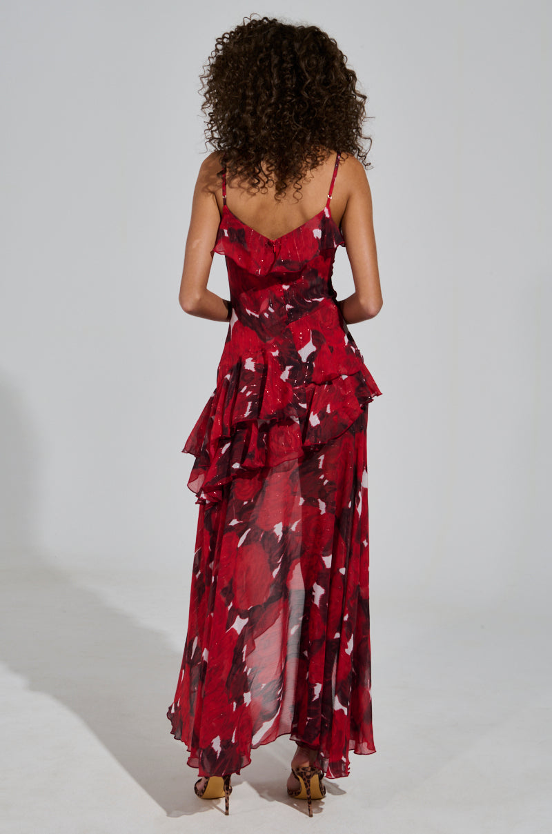 A person with curly hair, wearing the MAEVE PRINTED RUFFLE MAXI DRESS adorned in a red and black floral pattern, stands facing away. The floor-length gown, featuring adjustable tank straps and ruffled layers, pairs beautifully with their strappy heels against the white backdrop.