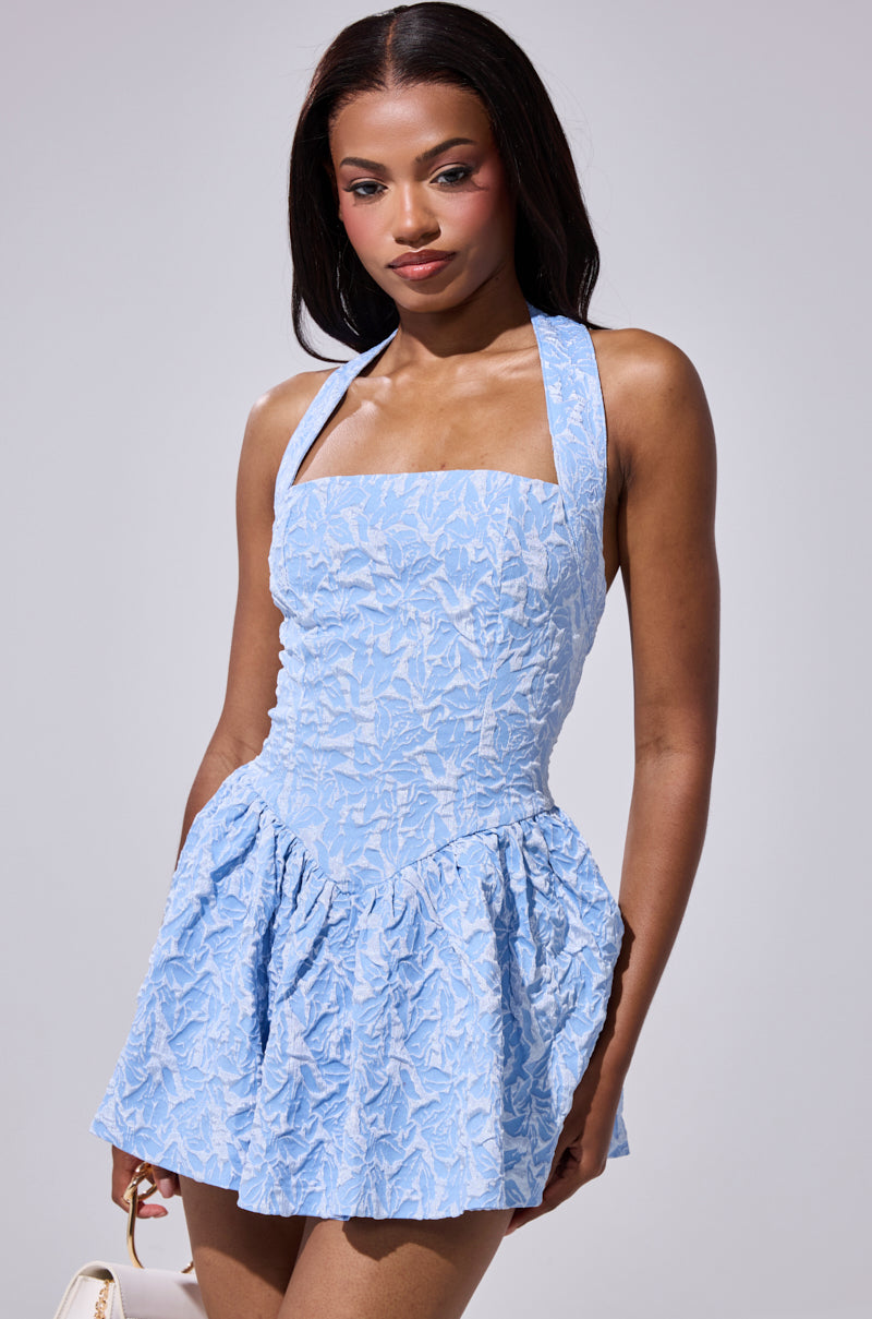 A woman with long dark hair stands against a neutral background wearing the LILY ANN TEXTURED MINI DRESS—a light blue skater dress with a fitted bodice and flared skirt—paired with a small, light-colored purse.