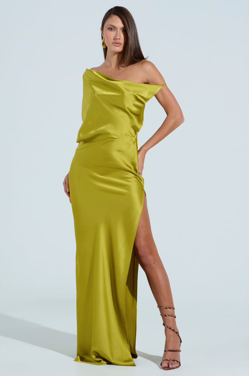 A woman confidently poses in a shiny chartreuse ISLA SATIN MAXI DRESS featuring a high slit and asymmetrical off-the-shoulder design. Her long brown hair flows down, and she completes the look with strappy black high heels against a solid, light background.