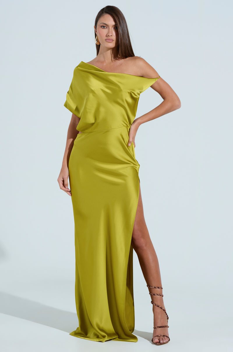 A woman stands confidently in an ISLA SATIN MAXI DRESS, a flowing chartreuse satin gown with a thigh-high slit. Her long brown hair cascades over one shoulder, and she dons strappy high heels against a soft, light blue background.