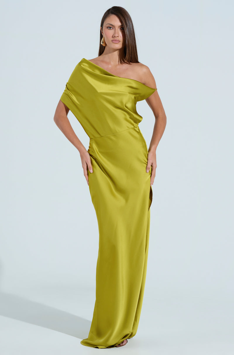 A woman poses confidently in an ISLA SATIN MAXI DRESS against a light blue background. The chartreuse dress features an asymmetrical off-the-shoulder neckline and a draped design. Her long brown hair complements her elegant hoop earrings.