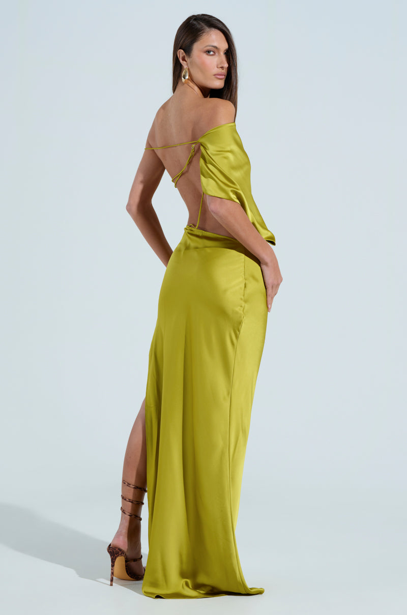 A woman confidently poses in the ISLA SATIN MAXI DRESS, showcasing its olive green, flowy fabric with a thigh-high slit and backless design. Her long dark hair accentuates hoop earrings and strappy heels against a light blue backdrop.