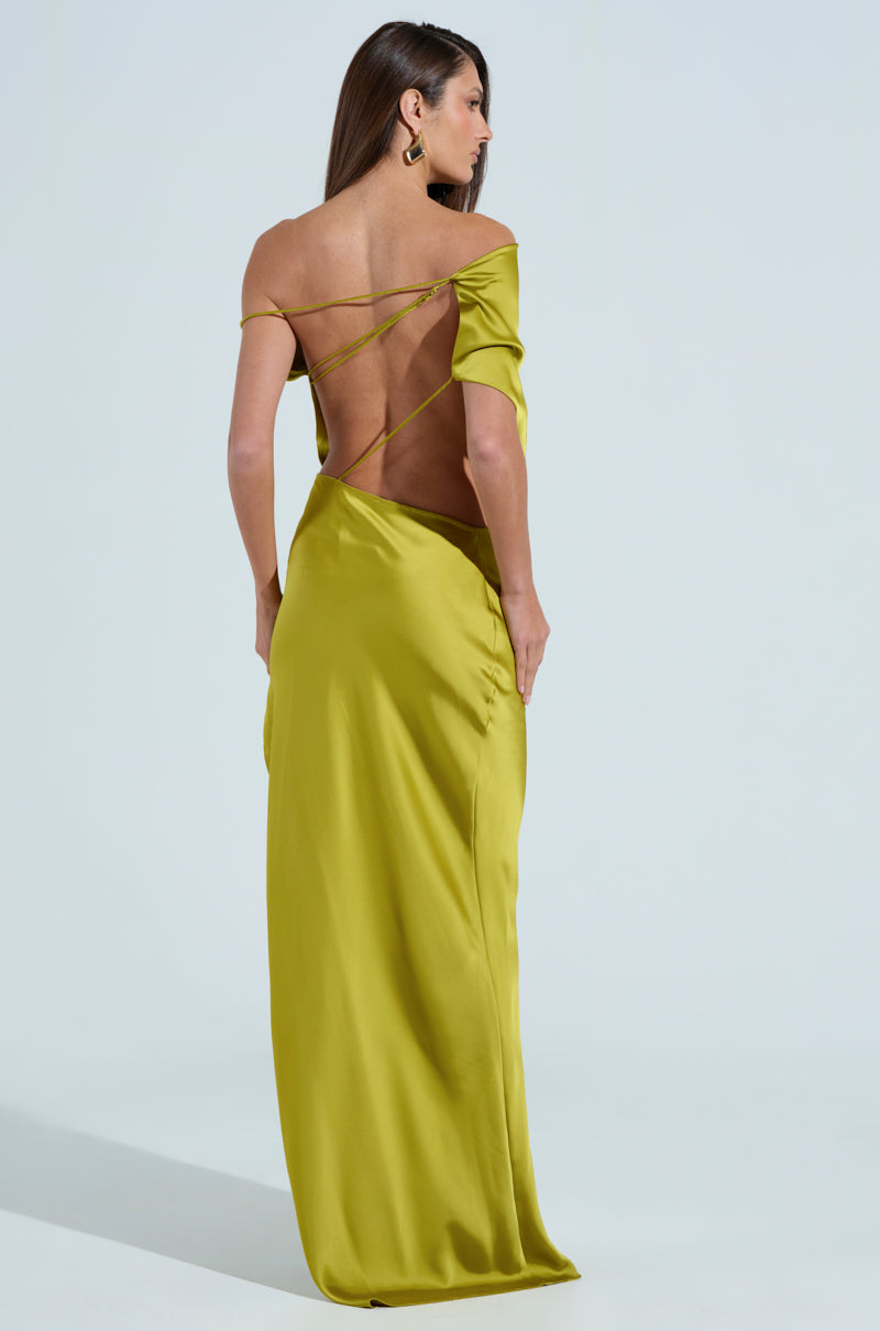 A woman with long hair wears the ISLA SATIN MAXI DRESS, a backless, asymmetrical off-the-shoulder chartreuse gown with thin straps crossing on her back as she stands against a plain, light-colored background.