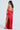 A woman poses in the SEXY CAN I SATIN MAXI DRESS, a backless red design featuring a cowl neck and thigh-high slit. She holds a gold clutch, wears strappy heels, and her long dark hair cascades down her back against a plain backdrop.