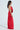 A person with long hair stands in profile against a plain background, wearing the SEXY CAN I SATIN MAXI DRESS. This red backless gown has a cowl neckline, draped front, smooth satin fabric, and a thigh-high side slit.