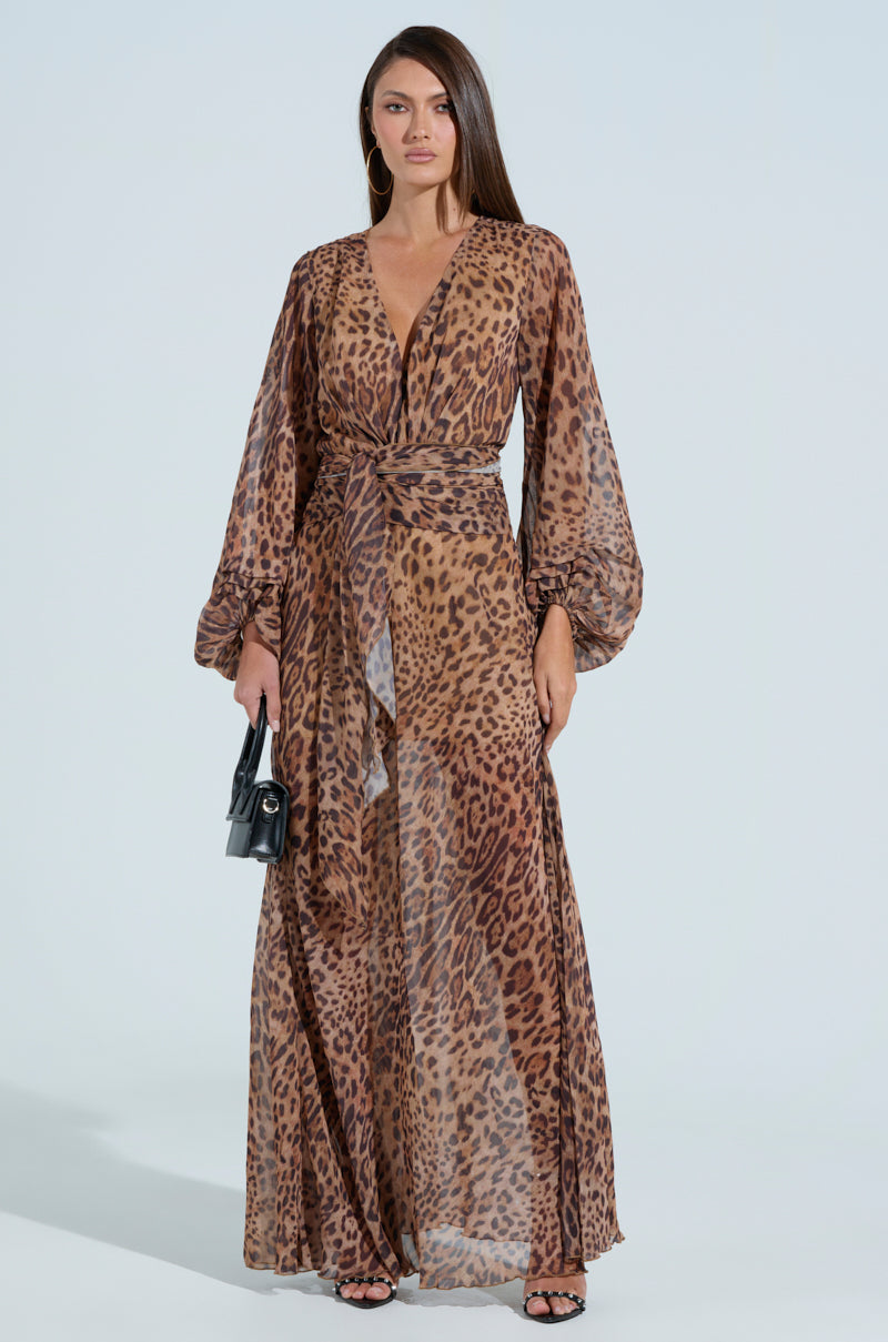 Against a light background, someone wears the RYAN LEOPARD PRINT MAXI DRESS featuring a plunging V-neckline and balloon sleeves. They carry a small black handbag and wear black strappy sandals.