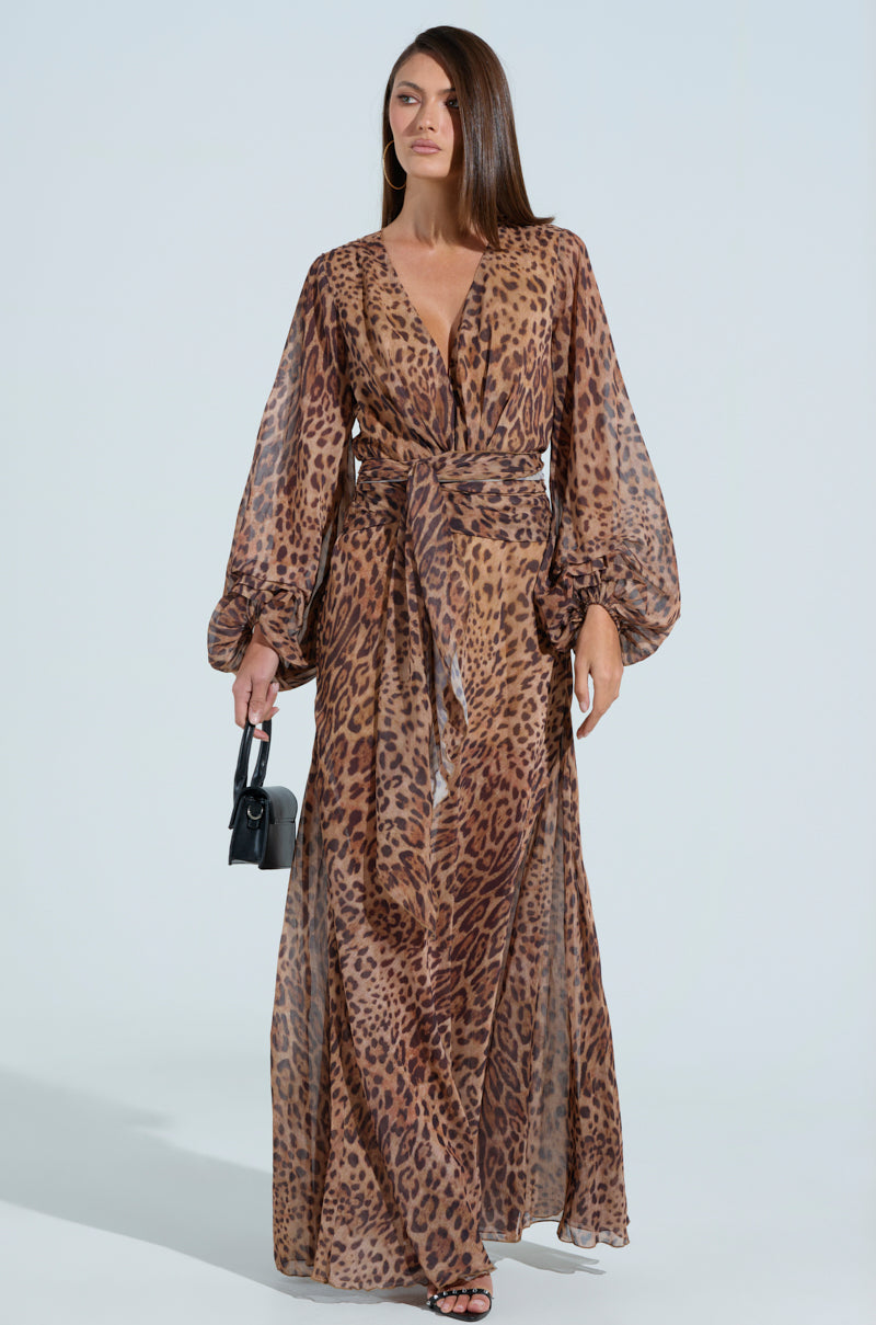 A person wearing the RYAN LEOPARD PRINT MAXI DRESS, featuring balloon sleeves and a plunging V-neckline, holds a small black handbag. Their long, straight hair and sandals stand out against a plain white background.