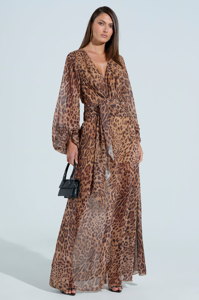A woman stands confidently in the RYAN LEOPARD PRINT MAXI DRESS, showcasing balloon sleeves and a plunging V-neckline. She carries a small black handbag, her long straight hair flowing against a simple, light-colored backdrop.