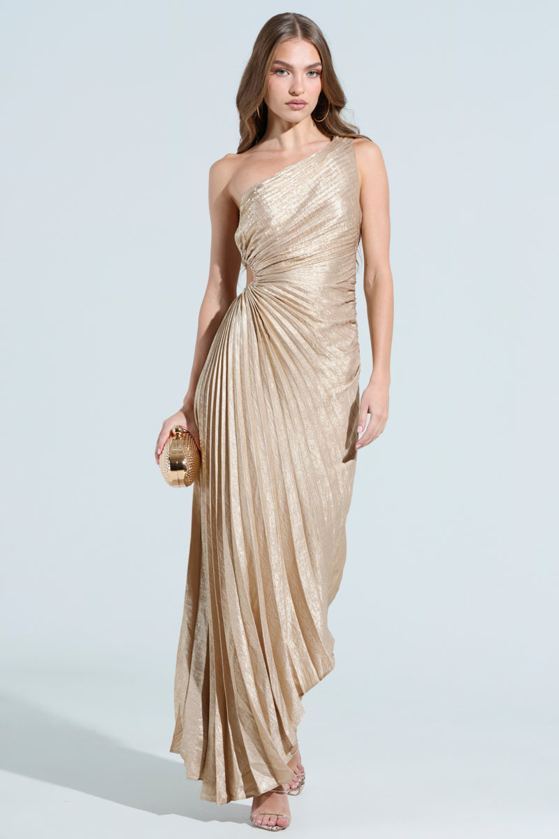 A woman confidently stands against a light backdrop in the JULIETTE METALLIC PLEATED MIDI DRESS, a shimmering gold one-shoulder gown with an asymmetrical hem. She holds a small gold clutch in her right hand, wears strappy heels, and her long hair flows loosely over her shoulder.