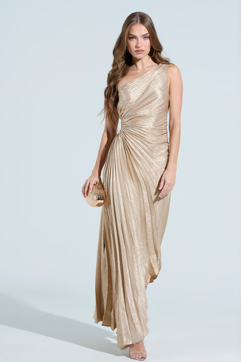 A woman in the elegant Juliette Metallic Pleated Midi Dress, featuring one shoulder and intricate detailing, holds a small, round clutch. Her long, wavy hair cascades as she stands against a simple light blue background.