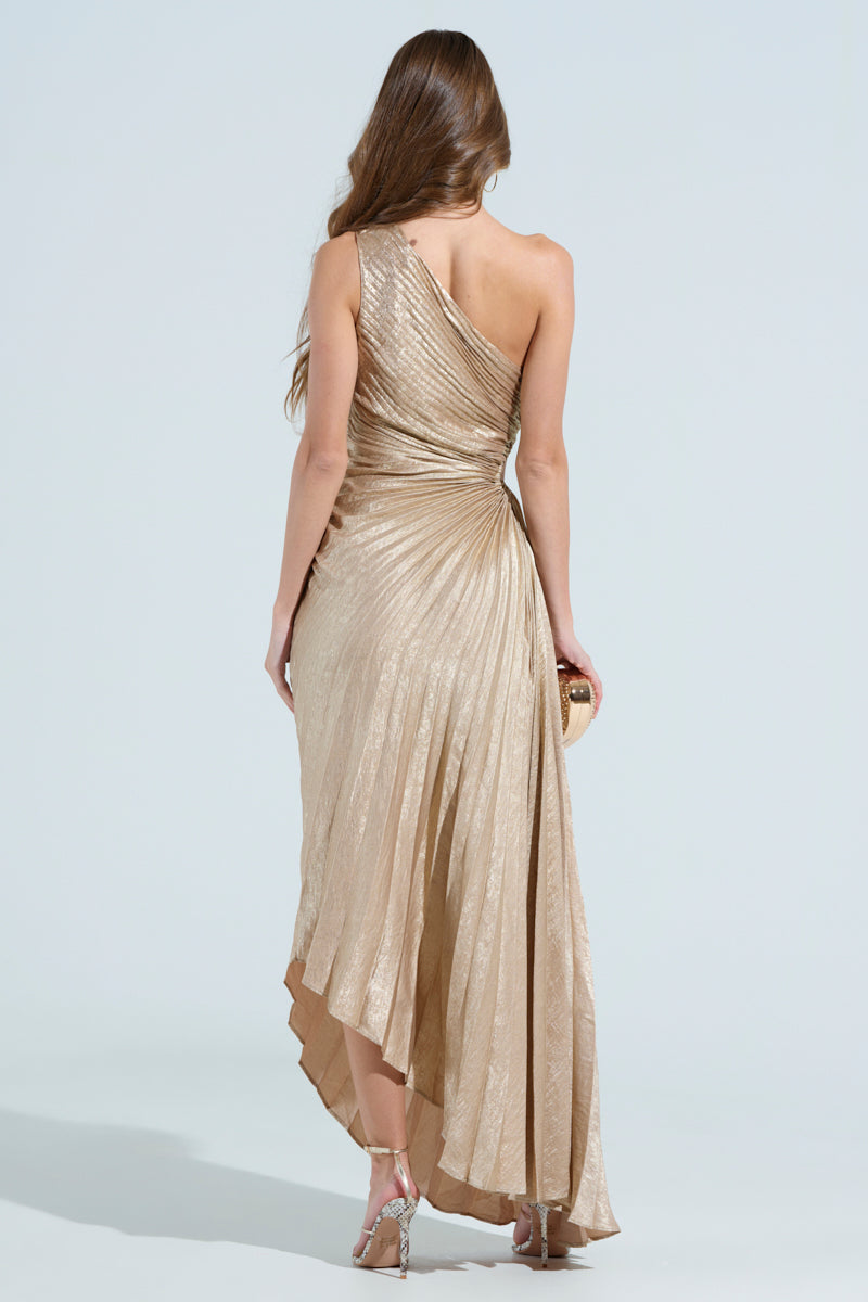 A woman with long hair is wearing a shimmering gold Juliette Metallic Pleated Midi Dress, holding a small gold clutch, and sporting silver high-heeled sandals against a neutral background.