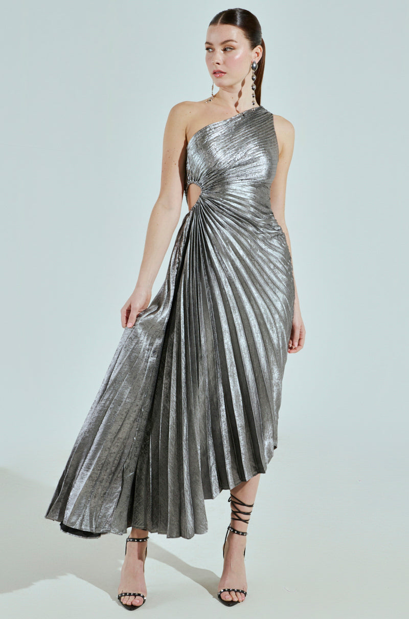A woman stands against a light background wearing the stunning "A LITTLE PARTY PLEATED MIDI DRESS IN SILVER," featuring an asymmetrical design, one-shoulder cut, and unique side detail. She completes her look with strappy black heels, large earrings, and elegantly pulled-back hair.