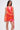 A woman stands against a white background wearing the ROXANNE PRINTED MINI DRESS in orange and pink, featuring a sleeveless halter style, open back, and asymmetrical hem. Her long straight hair flows as she looks to the side.