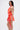 A woman with long straight hair wears the ROXANNE PRINTED MINI DRESS, a pink and orange halter mini dress with a tied waist and open back, standing sideways to reveal a tattoo on her upper arm against a plain white background.