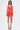 A woman with straight dark hair and visible leg tattoos wears the ROXANNE PRINTED MINI DRESS—an orange and pink wrap style with a v-neck, side tie, open back, and platform sandals, standing against a plain white background.