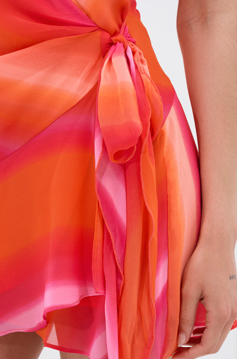 Close-up of a person wearing the ROXANNE PRINTED MINI DRESS, a flowing halter dress in bold orange, pink, and red with a side tie detail. Their hand rests by their side against a plain background. 6