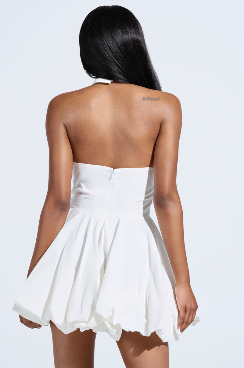 A woman with long black hair is seen from the back wearing the LORRAINE TIE FRONT MINI DRESS, a strapless white piece perfect for yacht parties. The dress fits at the top and flares into a pleated skirt, revealing a tattoo on her upper back against a plain, light background. 3