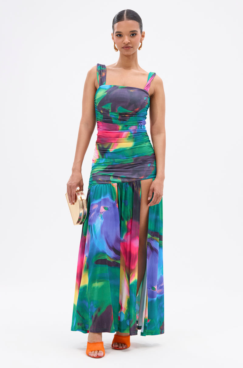 A woman stands against a white background wearing the ALAIA PRINTED MAXI DRESS in colorful jewel tones with a ruched floral pattern, paired with orange heels and a small gold clutch. 0