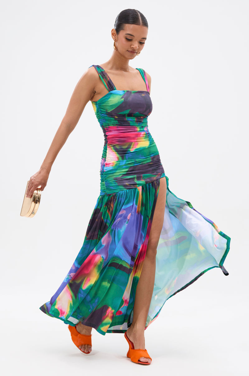 A woman walks gracefully against a white background in a jewel-toned, fitted ALAIA PRINTED MAXI DRESS with a high slit and flowing skirt, holding a gold clutch, wearing orange heeled sandals, and her hair tied back. 1