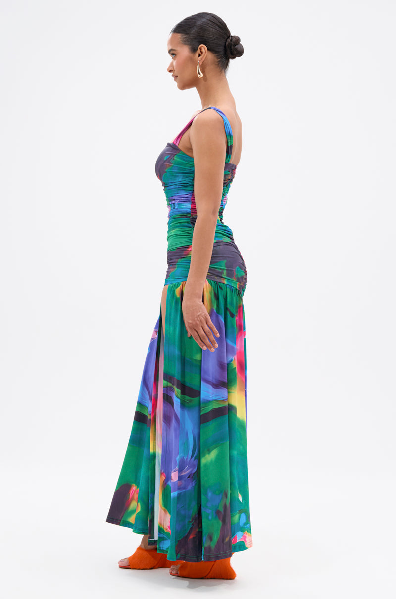 A woman in profile wears the ALAIA PRINTED MAXI DRESS, a fitted, sleeveless maxi with a ruched bodice and flowing skirt in jewel-tone green, blue, and purple. Her hair is in a bun; she pairs orange shoes with hoop earrings. 3