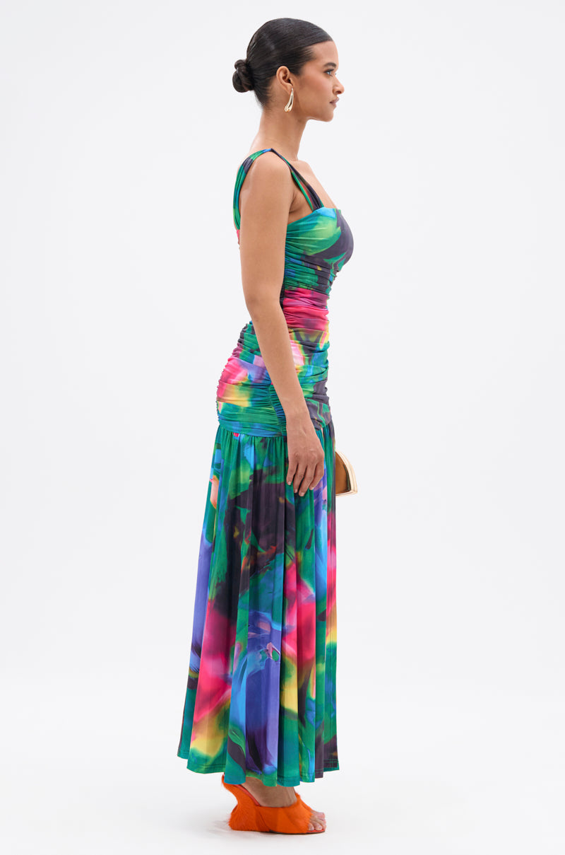 A woman stands in profile wearing an ALAIA PRINTED MAXI DRESS with a fitted bodice and flowing skirt. Her hair is in a bun, she wears hoop earrings and orange shoes, and holds a clutch against a plain white background. 4