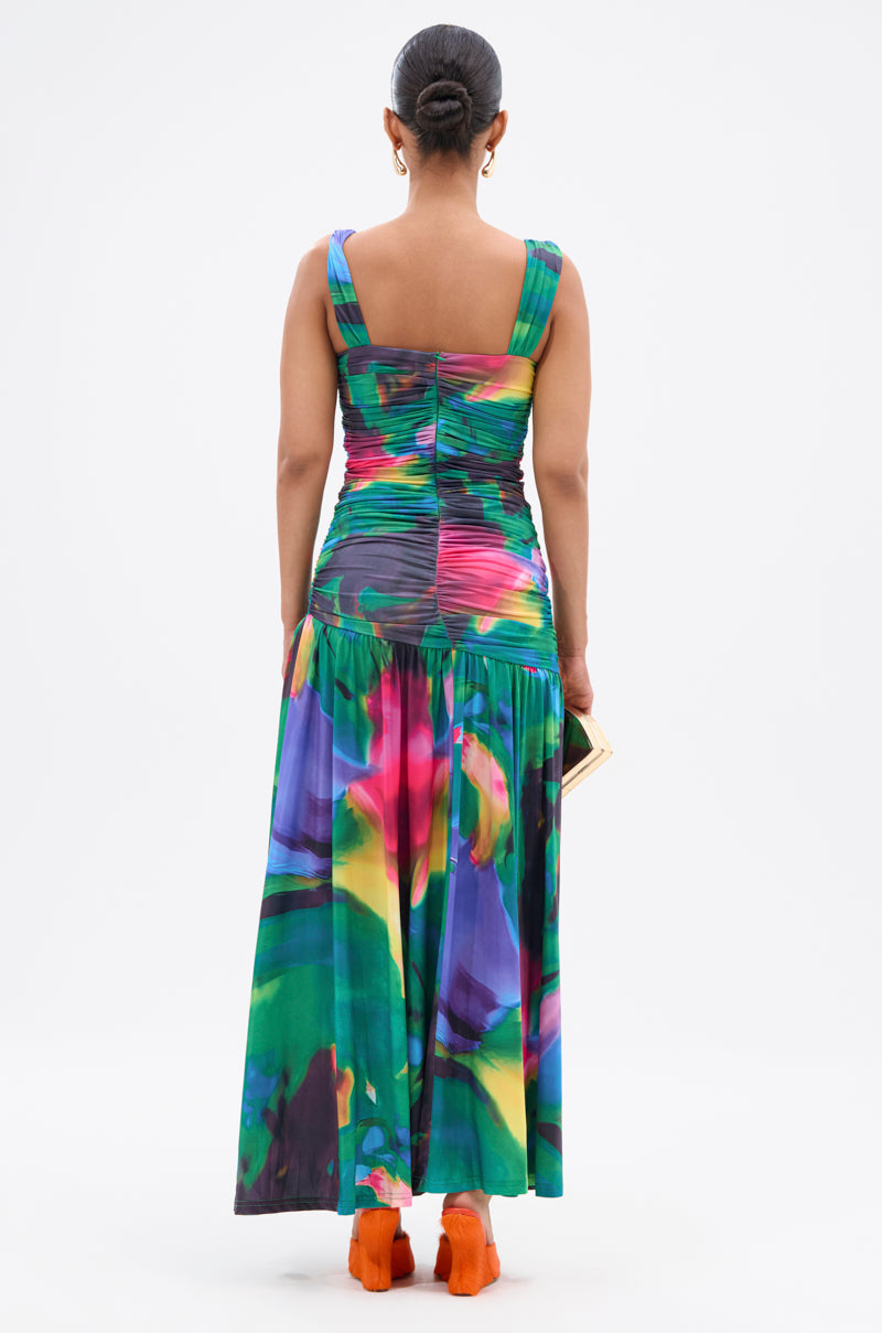 A woman with dark hair in a bun stands facing away in the ALAIA PRINTED MAXI DRESS, featuring multicolor abstract jewel tones, a long pleated skirt, bright orange shoes, and holding a small rectangular clutch. 5