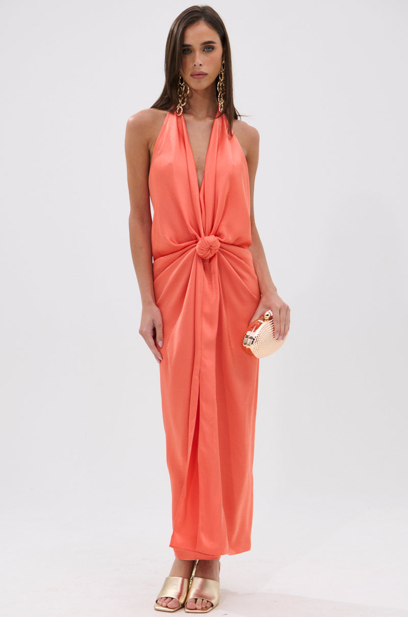 A woman models the KNOT MY PROBLEM SATIN MIDI DRESS—a sleeveless, halter-neck midi with waist knot detail—paired with a beige clutch, gold earrings, and open-toe heels against a white background. 0