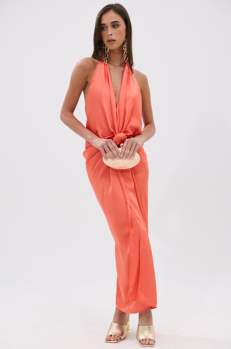 A woman stands against a white background wearing the KNOT MY PROBLEM SATIN MIDI DRESS, a sleeveless coral midi dress with a plunging halter neckline, paired with gold sandals, large gold earrings, and a small beaded clutch. 1