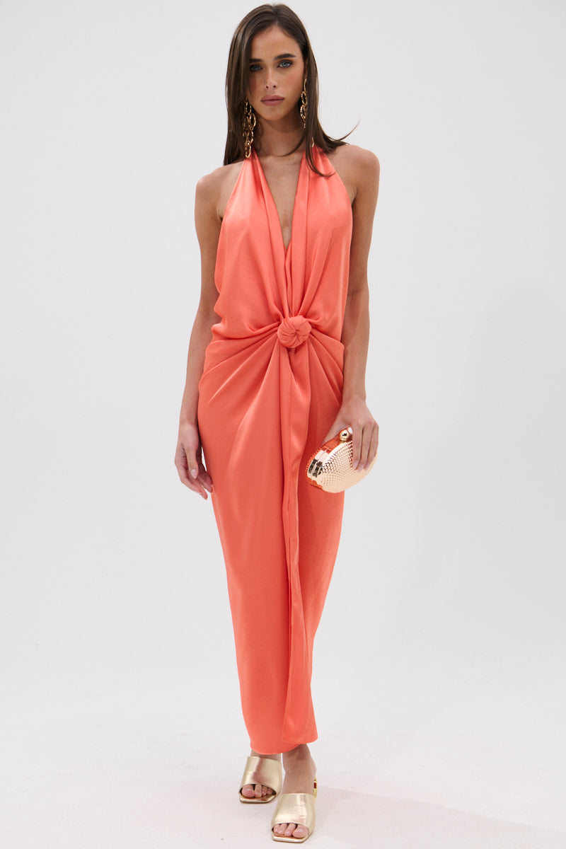 A woman stands against a white background wearing the KNOT MY PROBLEM SATIN MIDI DRESS—a sleeveless, satin midi dress with a plunging halter neckline and front knot—styled with gold earrings, sandals, and a small gold clutch. 2