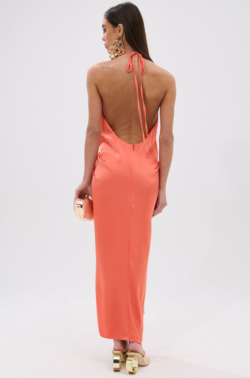 A woman models the KNOT MY PROBLEM SATIN MIDI DRESS in coral, featuring a backless cut and plunging halter neckline. She holds a gold clutch, wears gold earrings, heeled sandals, and has straight hair down against a white background. 5