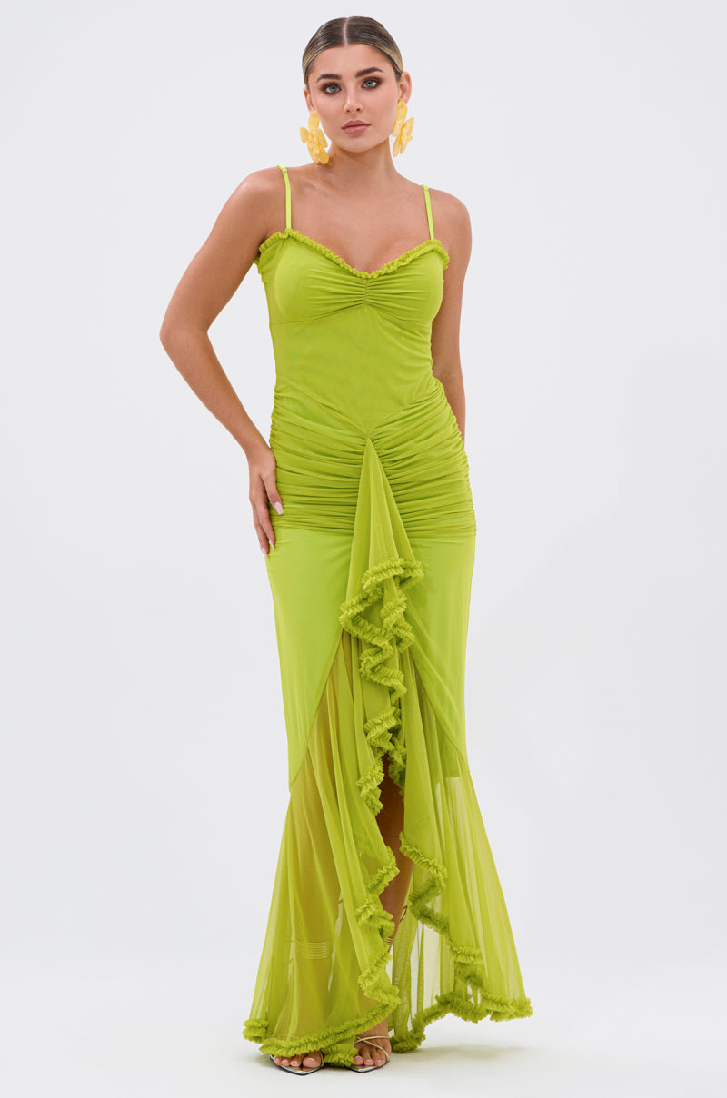A woman models the TATI RUFFLE MAXI DRESS IN LIME—featuring thin straps, ruched detailing, and a ruffle-trimmed V-neckline—against a plain white background, styled with slicked-back hair and bold yellow statement earrings.
