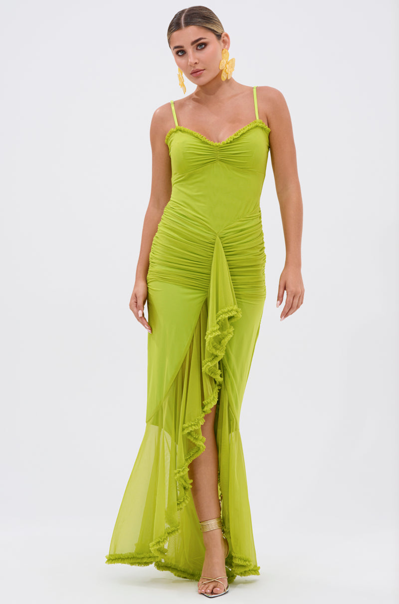 A woman in the TATI RUFFLE MAXI DRESS IN LIME, featuring ruched ruffle mesh details and a high slit, stands against a white background with her hair pulled back, accessorized with gold earrings and heels.