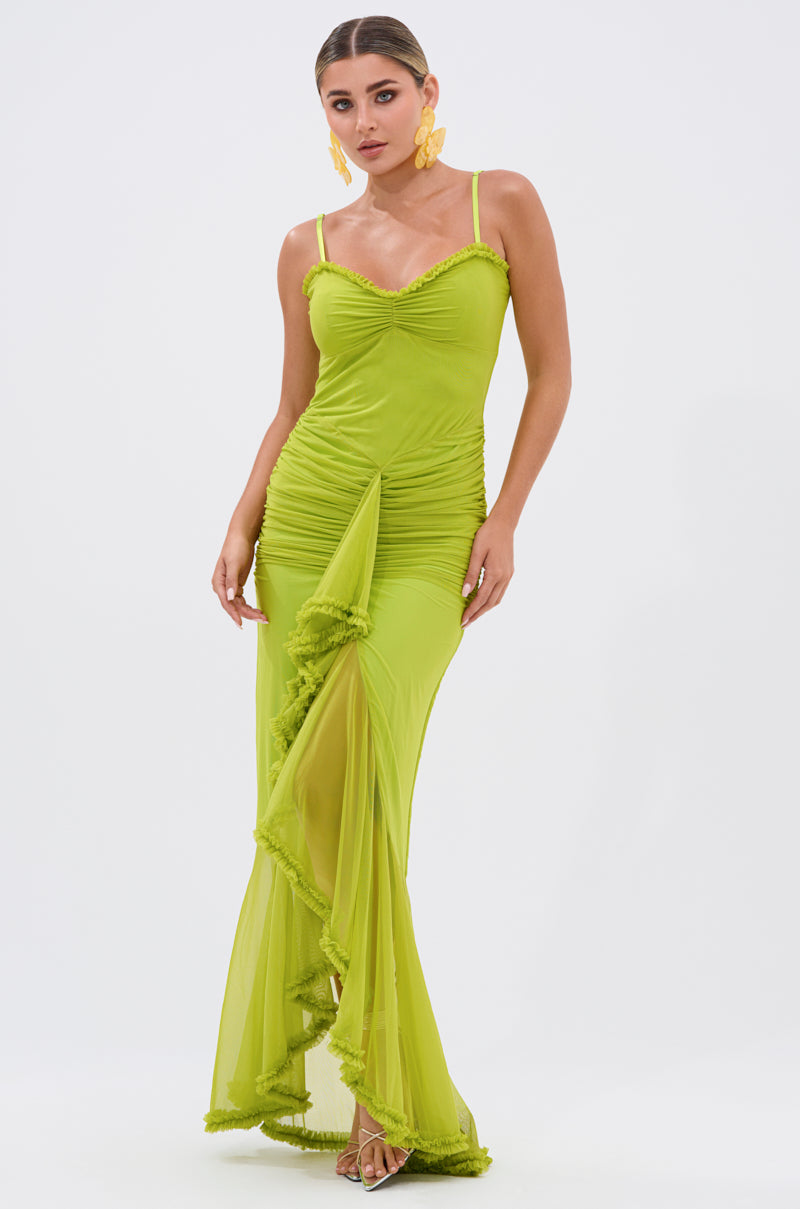 A woman wears the TATI RUFFLE MAXI DRESS IN LIME—featuring spaghetti straps, ruched details, and a ruffled asymmetrical sheer skirt—standing against a white background with slicked-back hair and statement earrings.