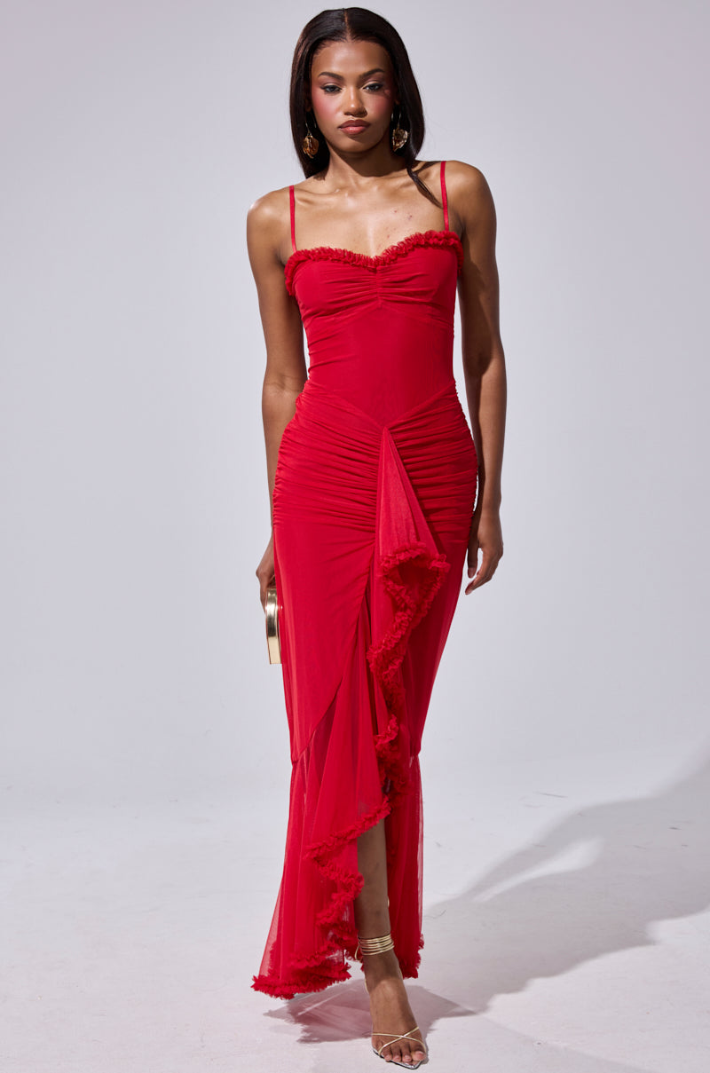 A woman in the TATI RUFFLE MAXI DRESS IN RED—featuring thin straps, ruching, and a ruffled hem—stands against a plain background, accessorizing with a clutch and strappy heels.