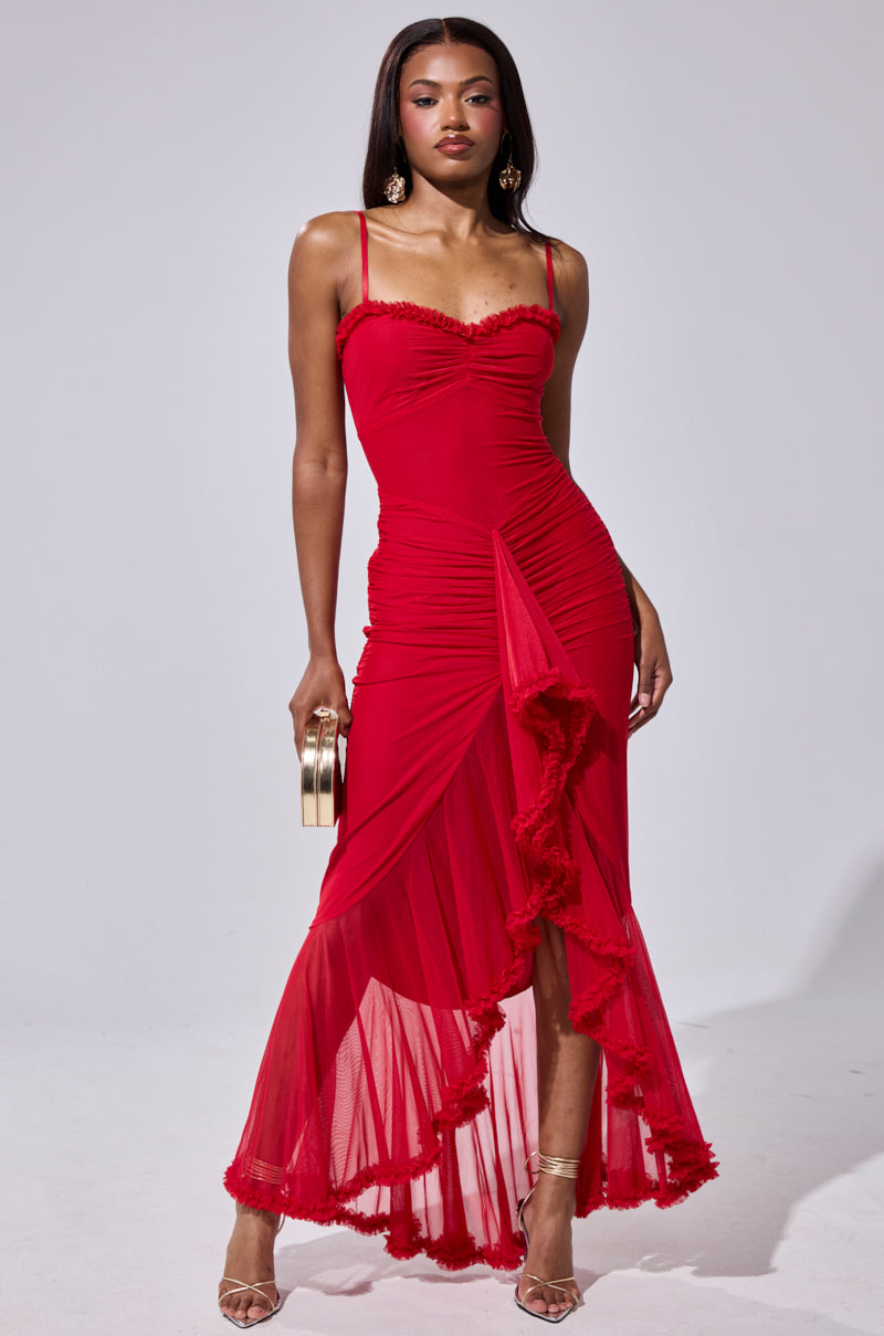 A woman stands confidently in the TATI RUFFLE MAXI DRESS IN RED, a sleeveless, fitted maxi with ruffle details and a sheer asymmetrical hem, accessorized with a gold clutch, strappy heels, and large hoop earrings.