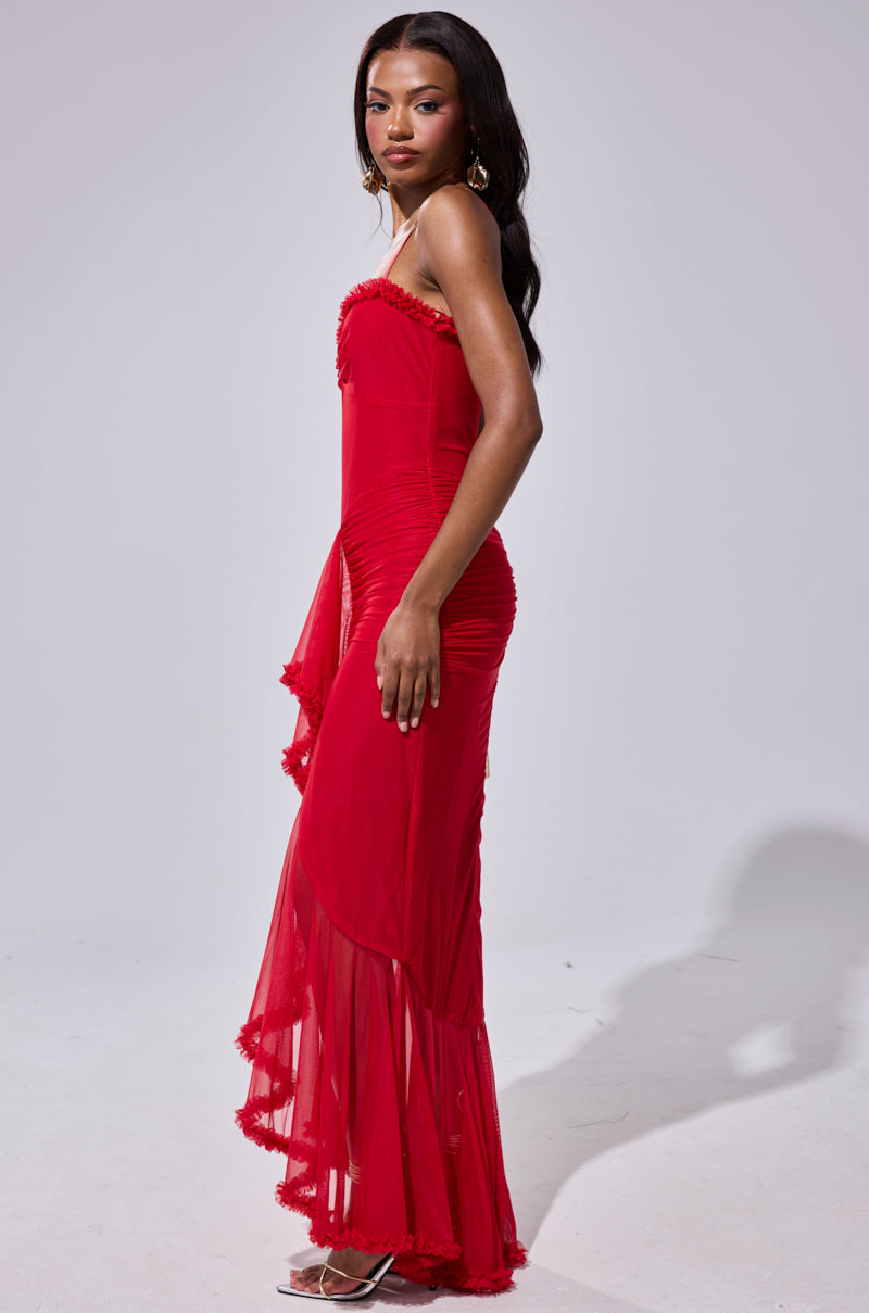 A woman with long dark hair, statement earrings, and strappy heels poses sideways in the TATI RUFFLE MAXI DRESS IN RED against a plain light background, looking at the camera.