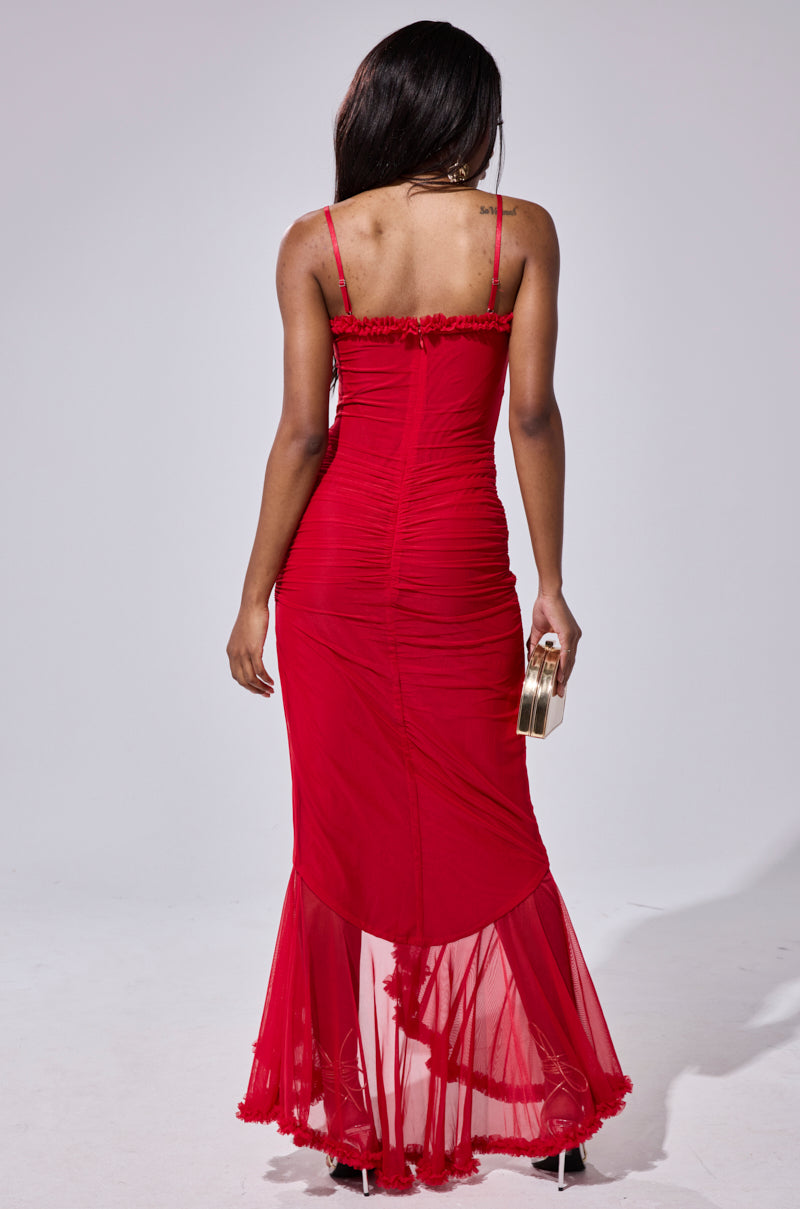 A woman with long dark hair wears the fitted TATI RUFFLE MAXI DRESS IN RED, featuring thin straps and a sheer hem. She stands with her back to the camera, holding a small gold clutch.