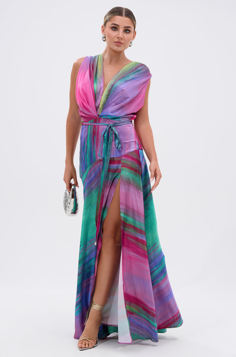 A woman wears the FOR HOLDING PRINTED MAXI DRESS—a sleeveless, floor-length dress with a pink, purple, green, and blue swirling pattern. It features a deep V neckline, belted waist, thigh-high slit. She completes the look with a clutch and heels.