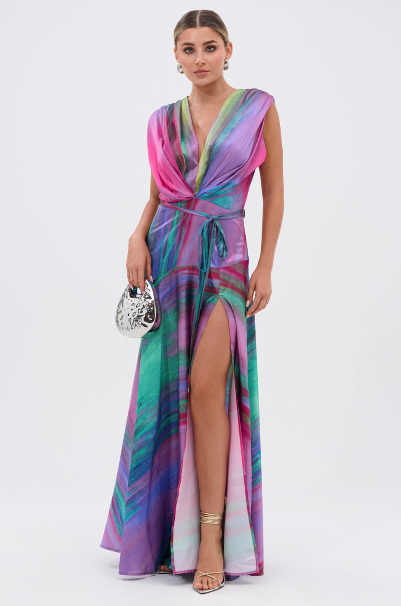 A woman models the FOR HOLDING PRINTED MAXI DRESS—a sleeveless, floor-length watercolor print dress with a thigh-high slit—paired with strappy heels and a small silver handbag, standing against a plain white background.