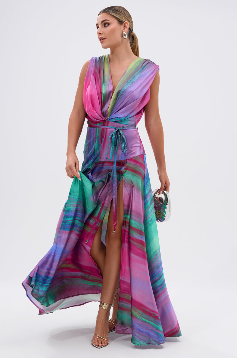 A woman wears the FOR HOLDING PRINTED MAXI DRESS with pink, purple, and green stripes on a white background. She carries a small handbag and pairs the dress with metallic high-heeled sandals.
