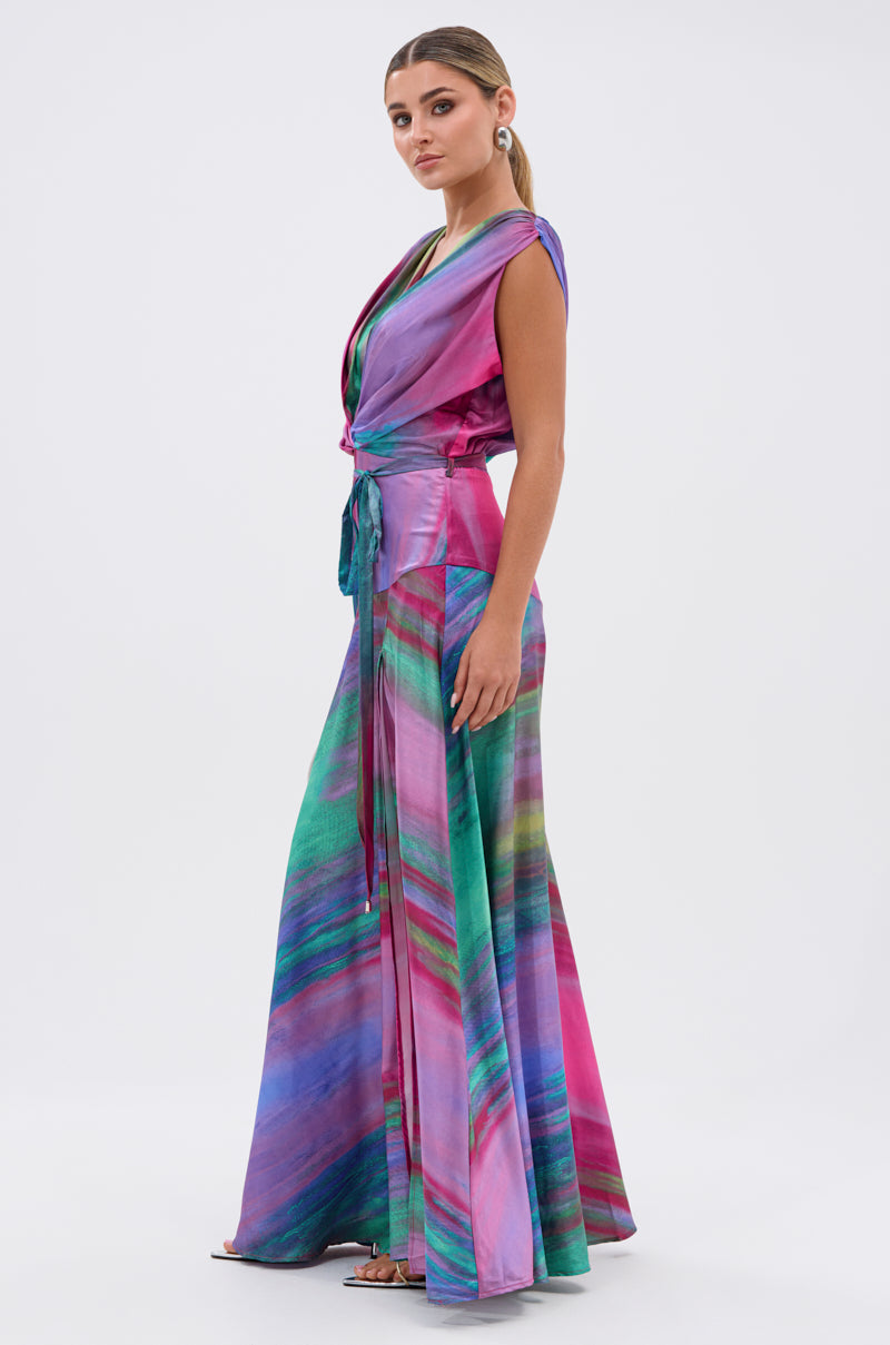 Woman poses in the FOR HOLDING PRINTED MAXI DRESS, a colorful sleeveless maxi with a wrap-style top, tie waist, and flowing skirt in vibrant purple, pink, blue, and green prints.