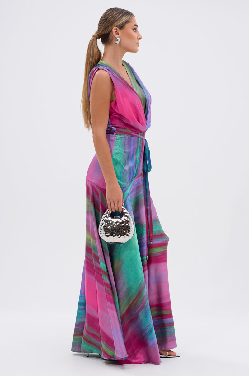A woman stands sideways wearing the FOR HOLDING PRINTED MAXI DRESS, featuring colorful pink, purple, and green patterns. Her long hair is tied back, and she carries a small white handbag with black embellishments.