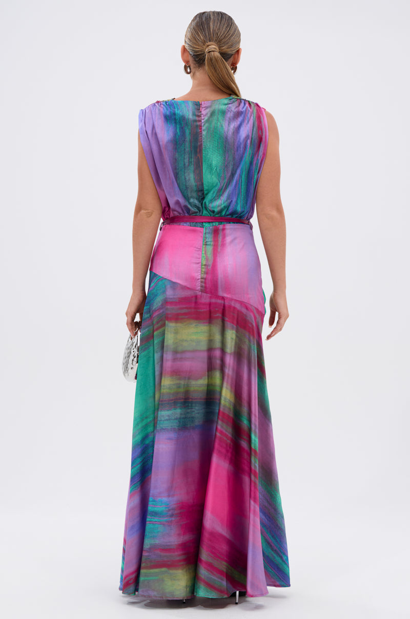 A woman with slicked-back hair stands facing away, wearing the FOR HOLDING PRINTED MAXI DRESS—a sleeveless satin maxi dress featuring a colorful abstract watercolor print in pink, green, and purple—while holding a small silver bag.