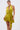A woman poses against a plain white background in the STAR OF THE SHOW SATIN MINI DRESS, a shiny chartreuse mini with a wrap-front bodice, ruffled hem, and side tie. Her hair is slicked back and she wears large gold earrings.