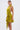 A woman wears the STAR OF THE SHOW SATIN MINI DRESS, a shiny chartreuse mini with a wrap-front bodice and large waist sash. She stands against a white background, her long hair tied back and bold gold earrings as accessories.